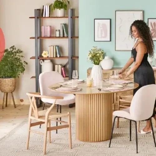 Woman setting a table in a stylish dining room with decor and plants.