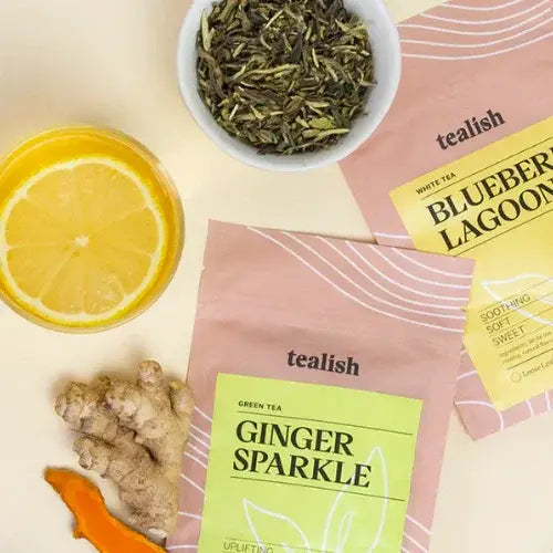 Tealish tea packages with ginger, lemon, and a bowl of green tea leaves on a light background