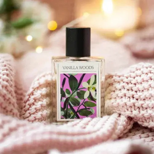 Vanilla Woods perfume bottle with pink label featuring green leaves on a soft pink fabric background.