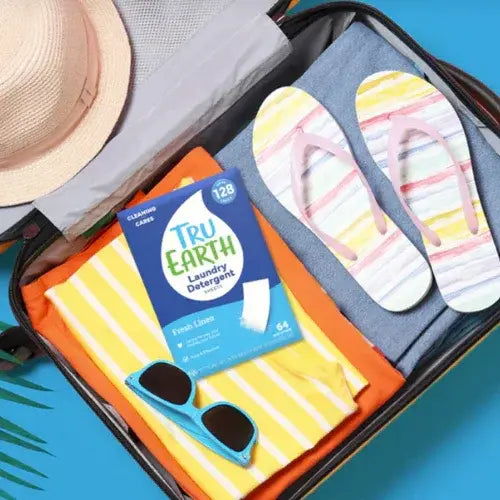 Open suitcase with colorful striped flip-flops, sunglasses, and a package of Tru Earth laundry detergent on a blue background.