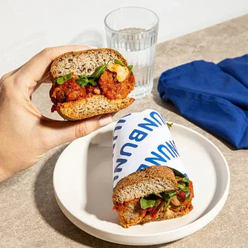 Person holding a sandwich with a glass of water and blue napkin on a neutral background