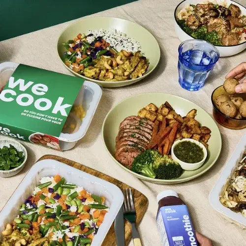 An assortment of healthy prepared meals from WeCook displayed on a table, including dishes with grilled meats, vegetables, rice, sweet potato fries, and cashews. A smoothie bottle, a container of cookies, and fresh herbs accompany the meals, creating a vibrant and nutritious spread.