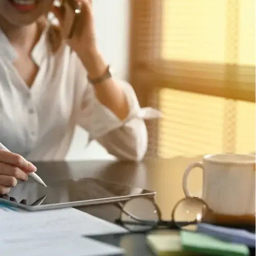 Businesswoman working on tablet while talking on phone at desk with coffee and documents — modern office productivity scene.
