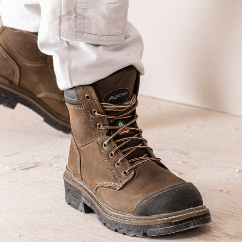 Brown work boot with black sole worn by a person on a light wooden floor.