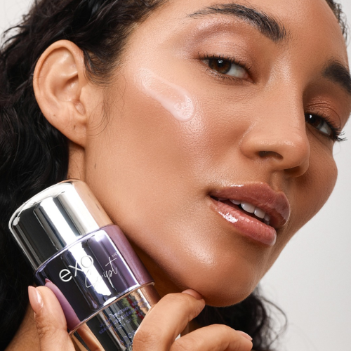 Woman applying skincare with a brush to her face, holding a makeup product.