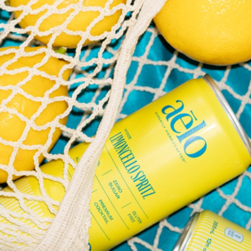 Yellow aēlo Lemoncella Spritz cans arranged on a turquoise surface with a white mesh net and lemons.