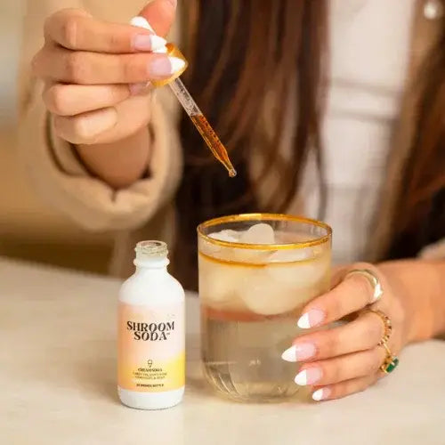 Person adding a drop of liquid from a dropper into a glass with 'Shroom Soda' bottle nearby.