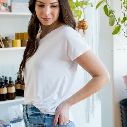 Woman wearing a white t-shirt and blue jeans in a room with shelves and plants.