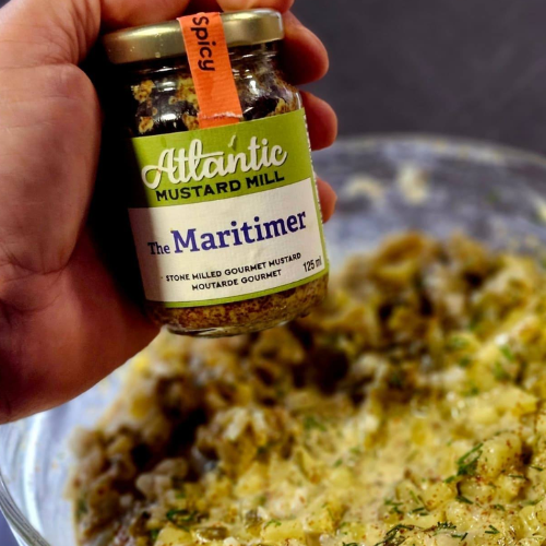 Hand holding a jar of Atlantic Mustard Mill 'The Maritimer' mustard with food in the background.