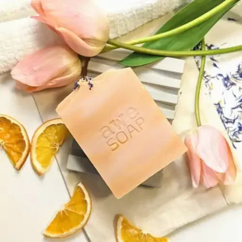 A handcrafted peach-toned bar of soap stamped with 'awe SOAP' resting on a soap dish, surrounded by soft pink tulips, dried orange slices, a white towel, and delicate blue flower petals. The arrangement is styled for a fresh, natural, and spa-like aesthetic.