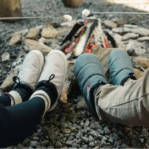 Two people sitting by a campfire, wearing cozy insulated slippers and thick socks, with marshmallows roasting over the flames. The ground is covered in gravel, and the fire is surrounded by large stones, creating a warm and rustic camping scene.