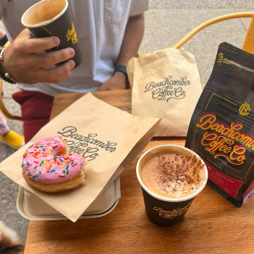 Two cups of coffee and a donut with 'Beachcomber Coffee Co' branding on a wooden table.