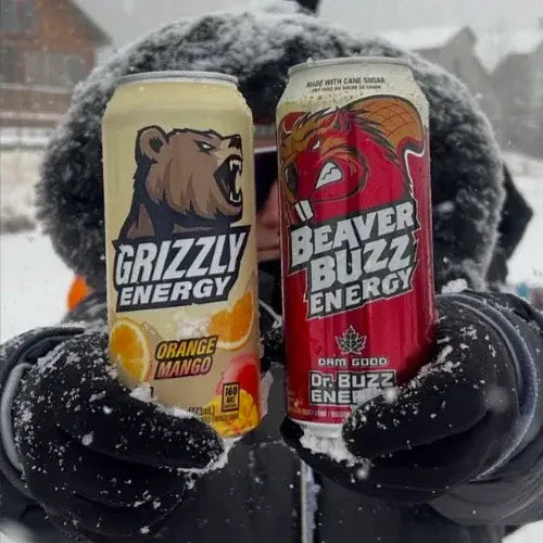 Two energy drink cans, 'Grizzly Energy' and 'Beaver Buzz Energy', held by a person in a snowy setting.