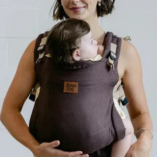 A woman wearing a dark brown baby carrier holds a baby close to her chest. The baby looks up at her while comfortably nestled in the carrier, which features adjustable beige straps and a leather patch that reads 'Beluga Baby.' The background is minimal with a light-coloured wall.