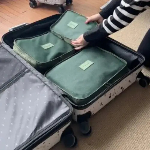 Person organizing a white rolling suitcase with green packing cubes labeled for travel essentials, placing them neatly inside on a wooden floor with a rug nearby.