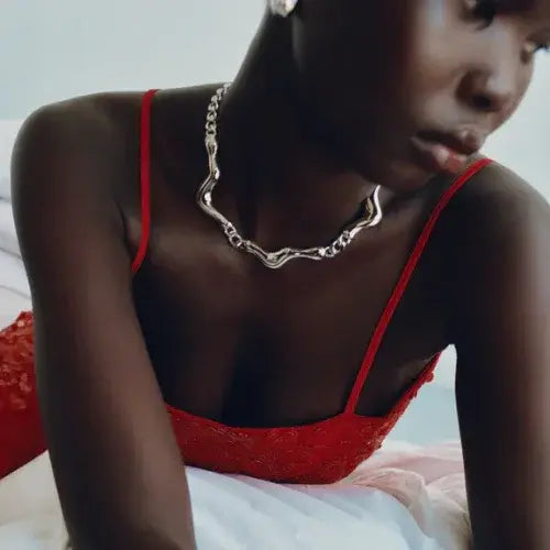 Close-up of a woman wearing a bold silver chain necklace and a red lace spaghetti strap dress. Her pose is elegant and slightly turned away, highlighting the jewellery and textured fabric against a soft, minimal background.