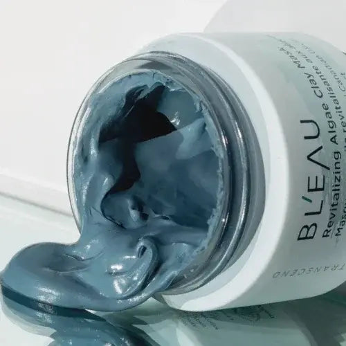 Open jar of BLEAU Revitalizing Algae Clay Mask with smooth blue formula spilling out, natural skincare product on reflective surface