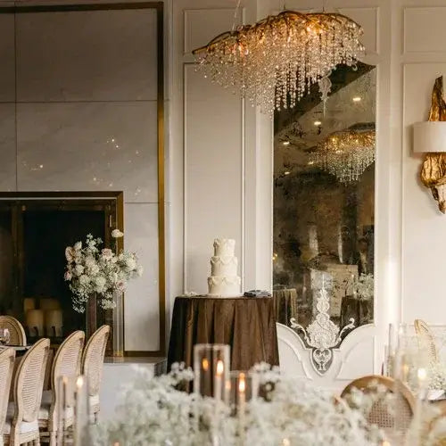 An elegant wedding reception setup featuring a tall, white tiered cake on a round table with a dark tablecloth. The room is adorned with a crystal chandelier, floral arrangements, candlelit tables, and luxurious gold and cream decor.