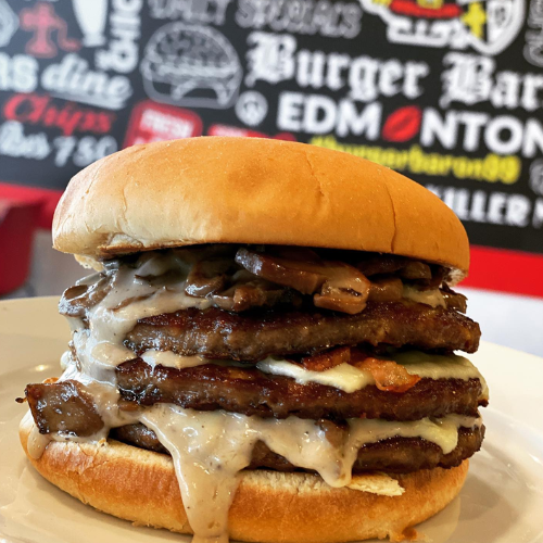 Stacked burger with multiple layers of meat and cheese on a bun, with a blurred background featuring text and logos.