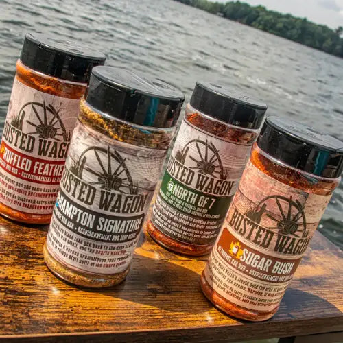 Four jars of Busted Wagon seasoning on a wooden surface with a water background