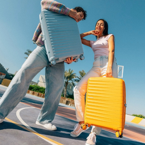 Two people outdoors under a clear blue sky, each holding a hard-shell spinner suitcase. One suitcase is light blue and is held by a person in a plaid shirt and jeans, while the other is bright yellow and is held by a person in a light pink top and white pants.