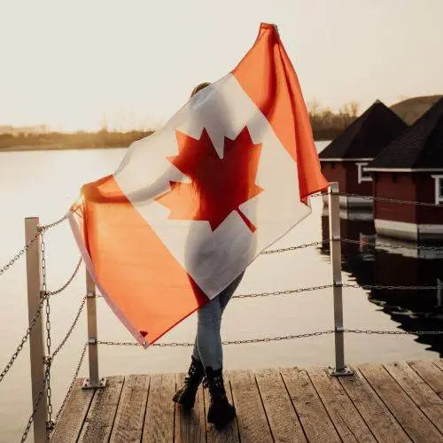 Person holding a Canadian flag on a dock by a body of water with red buildings in the background.