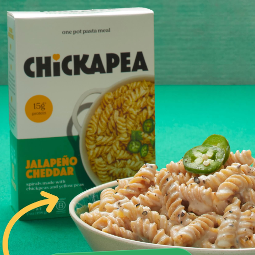 A box of Chickapea Jalapeño Cheddar one pot pasta meal stands behind a bowl of prepared spiral pasta with sliced jalapeño on a bright teal background.