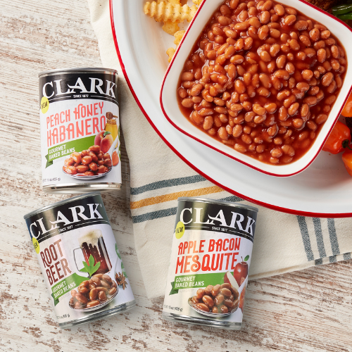 Three cans of Clark baked beans on a wooden surface with a bowl of beans and cornbread in the background.