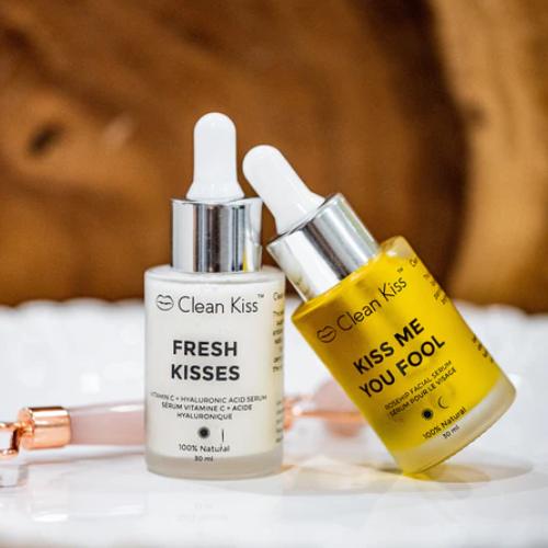 Two skincare serum bottles labelled 'Fresh Kisses' and 'Kiss Me You Fool' on a white surface with a brown background.