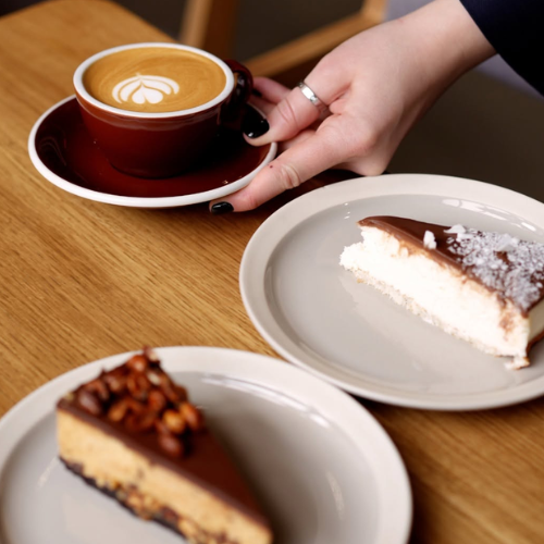 A hand with black nail polish and a silver ring holds a dark red ceramic cup of latte art coffee on a matching saucer, set on a wooden table next to two plates of dessert.