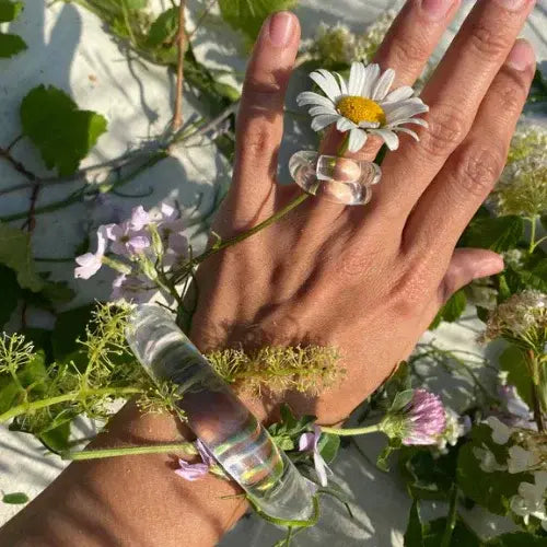 Hand wearing clear lucite bracelet and ring surrounded by wildflowers – Playful, sculptural jewellery from Canadian designer Corey Moranis