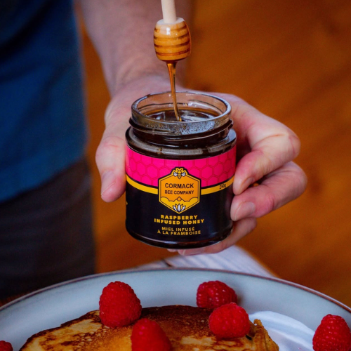 Person drizzling honey from a jar labelled 'Cormack Bee Company' onto pancakes with raspberries.