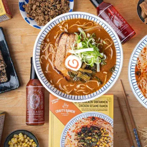 Bowl of ramen with garnishes on a wooden table with a Crafty Ramen box and hot sauce.