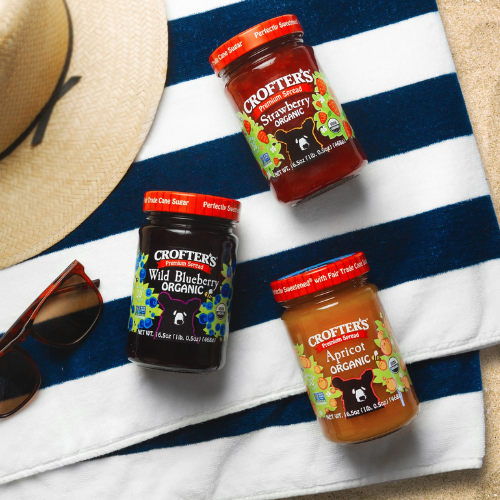 Three jars of Crofter's Organic jams on a blue and white striped towel with a hat and sunglasses.