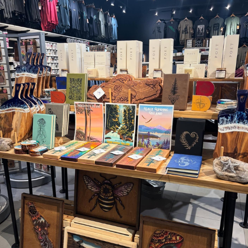 A retail display table showcasing a variety of handcrafted wooden art pieces, including laser-engraved wood panels, a carved wooden horse, and colorful framed prints, set inside a store.