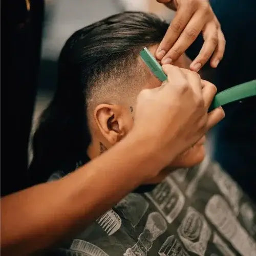 A close-up of a person getting a precision haircut at a barbershop. A barber uses a straight razor to shape the client's hairline while the client wears a printed cape featuring hair clipper illustrations.