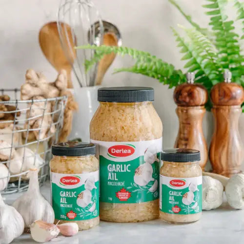 Three jars of Derlea Garlic Oil on a kitchen counter with garlic and cooking utensils in the background.
