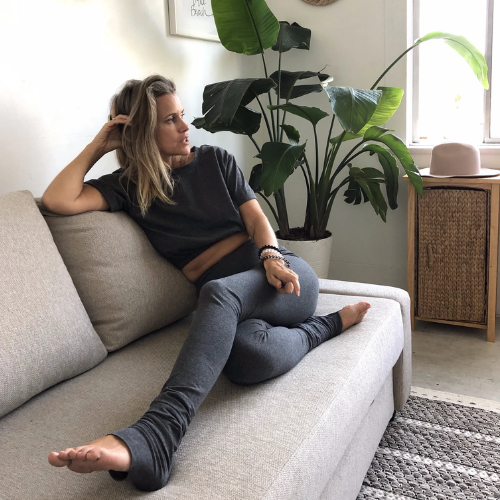 A woman lounges on a light gray fabric sofa in a bright, modern room. She is wearing a dark charcoal gray t-shirt and matching dark gray leggings.