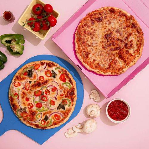 Two pizzas on blue and pink boards with ingredients on a pink background