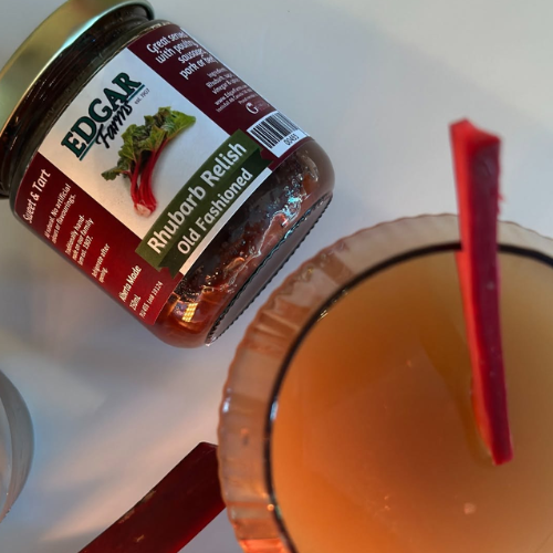 A jar of Edgar Farms Rhubarb Relish shown on a white surface next to a glass of amber-colored drink garnished with a rhubarb stick.