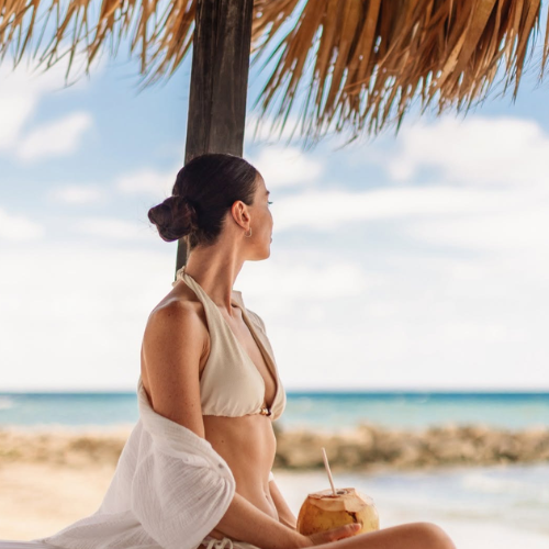 Woman sitting on beach looking out at water with coconut drink in her hand