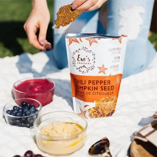 Eve's Crackers chili pepper pumpkin seed cracker being held over a container of crackers with a picnic setting.