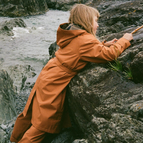 Person in an orange coat standing on rocks by a stream