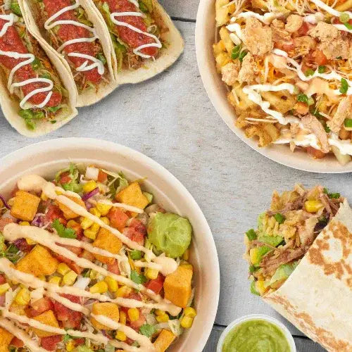 A flat lay of vibrant Mexican-inspired dishes, including soft shell tacos topped with salsa and crema, a burrito bowl with corn, guacamole, and crispy toppings, a plate of loaded nachos or chilaquiles, and a partially wrapped burrito with shredded meat. A small container of green salsa sits nearby on a light wooden surface.