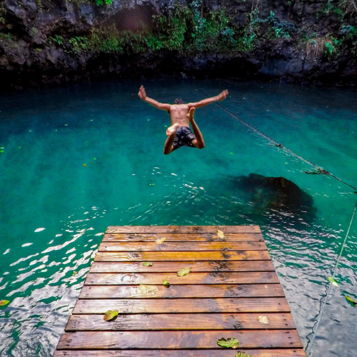 Person jumping off a wooden platform into clear blue water with a rocky cliff in the background