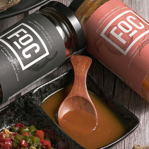 Two bottles of FOC sauce with a wooden spoon in a bowl on a wooden surface.