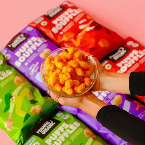 Hand holding a glass bowl of popcorn with Puffs Soufflé and Puffles packages in the background.