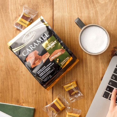 Packaged caramel snacks on a wooden table with a laptop and cup of coffee.