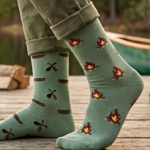 A close-up of a person's feet wearing sage green crew socks with a pattern of campfires and camp tools, paired with rolled-up olive green pants, set against a rustic outdoor background.