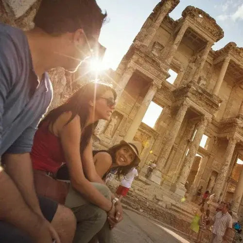 Three friends sitting and smiling in front of the ancient ruins of the Library of Celsus in Ephesus, Turkey, as the sun shines brightly in the background. The historic stone structure and scattered tourists create a vibrant travel atmosphere.